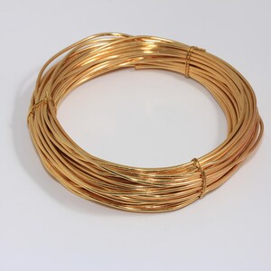 Puede incluir: Una bobina de alambre de color dorado, bien enrollada y asegurada. El alambre es de un dorado metálico brillante, y la bobina está centrada en una superficie blanca. Una regla rosa está en el fondo.