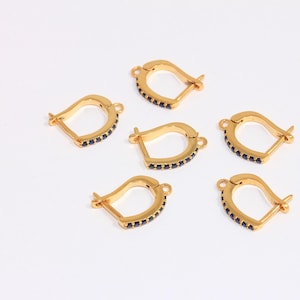 May include: Six gold-colored earring clasps with a curved design. Each clasp features a row of small, dark blue stones along the inner curve. The clasps are arranged on a white surface.