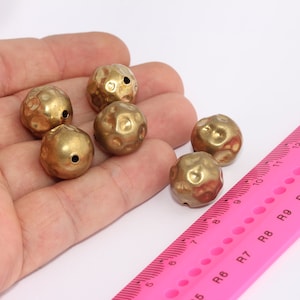 May include: Five gold-colored metal beads with a hammered texture and a hole in the center. The beads are arranged in a hand, with a pink ruler in the background.
