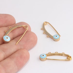May include: Three gold safety pins with blue evil eye charms. The charms have a white center and a blue enamel finish.