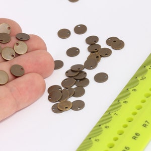 以下が含まれることがあります： 手の中に、小さな丸いアンティークゴールドの金属製のチャームがいくつか置かれています。チャームには中央に穴が開いています。背景には黒い目盛りがついた緑色の定規があります。