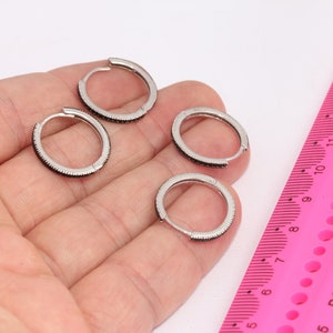 May include: Four silver hoop earrings with black accents. The earrings are displayed on a hand against a pink ruler.