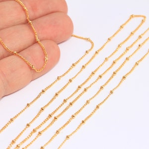 May include: A close-up of a gold beaded chain. The chain is laid out on a white surface and is shown in multiple lengths.