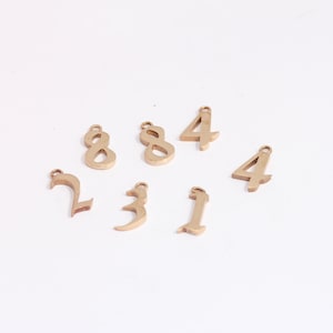 May include: A set of gold-colored metal number charms, including the numbers 1, 2, 3, 4, and 8. The charms are arranged on a white surface.