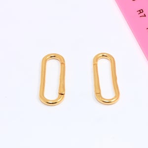 May include: Two gold-coloured, oval-shaped clasps are shown against a white background. The clasps are positioned side-by-side. A pink ruler is partially visible in the background, providing a size reference.