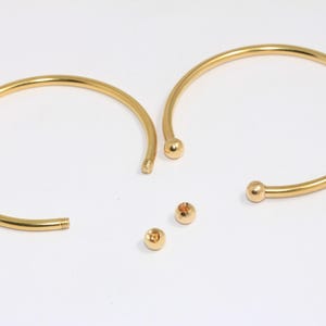 Può includere: Due braccialetti dorati con una palla ad ogni estremità. I braccialetti sono aperti e le palle sono separate.