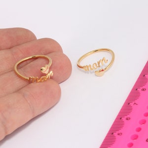May include: Two gold rings with the word "mam" spelled out on each ring. The rings are open and have a small heart charm on the end of the word.