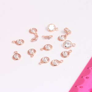Könnte beinhalten: Eine Sammlung von 14 roségoldfarbenen Metall-Anhängern mit klaren Kristall-Akzenten. Jeder Anhänger hat einen runden Kristall mit facettiertem Design und eine kleine Schlaufe zum Anbringen an Schmuck.
