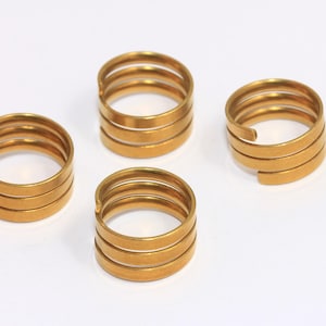 Puede incluir: Cuatro anillos de metal de color dorado, cada uno con tres espirales. Los anillos están dispuestos en semicírculo sobre una superficie blanca.