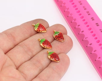 Puede incluir: Cuatro abalorios de color dorado en forma de fresas con esmalte rojo y verde. Los abalorios están en una mano con una regla rosa en el fondo.