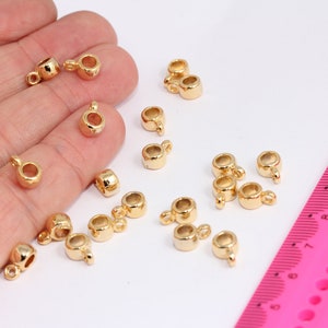 Puede incluir: Primer plano de un grupo de tapas de cuentas de metal de color dorado con un bucle en un extremo. Las tapas de cuentas están dispersas sobre una superficie blanca con una regla rosa en el fondo.