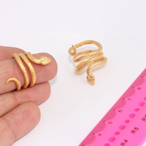 Könnte beinhalten: Zwei goldfarbene Schlangenringe. Der Ring links wird an einem Finger getragen. Der Ring rechts liegt flach auf einer weißen Oberfläche. Ein rosa Lineal befindet sich im Hintergrund.