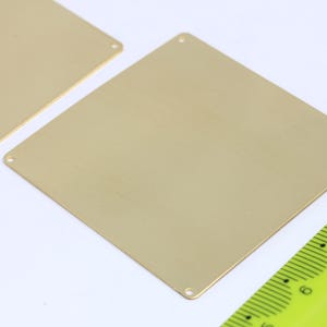 Puede incluir: Dos placas de metal de color dorado con esquinas redondeadas y agujeros en las esquinas. Las placas están sobre una superficie blanca.