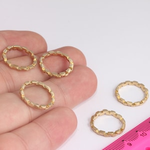 以下が含まれることがあります： 小さな正方形のリンクでできた、金色の金属製のリングが4つ。リングは手に置かれており、背景にはピンク色の定規がある。