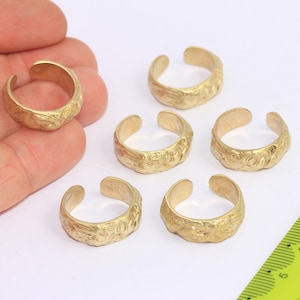 May include: Six gold-toned adjustable rings with a textured surface. The rings are displayed on a white surface with a ruler in the background.