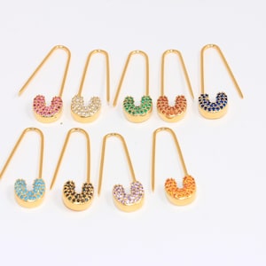 May include: Ten gold safety pin earrings with colorful rhinestones. The earrings are arranged in two rows of five. The colors of the rhinestones are pink, white, green, orange, blue, turquoise, black, white, orange, and red.