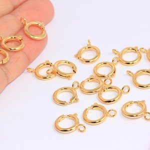 Può includere: Un gruppo di 12 chiusure a moschettone in metallo dorato, un tipo di minuteria per gioielli, sono sparsi su una superficie bianca. Un righello rosa è sullo sfondo.