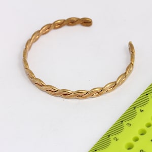 May include: A gold-colored, open cuff bracelet with a braided design. The bracelet is shown on a white surface next to a green ruler. The bracelet's diameter is approximately 2.5 inches.