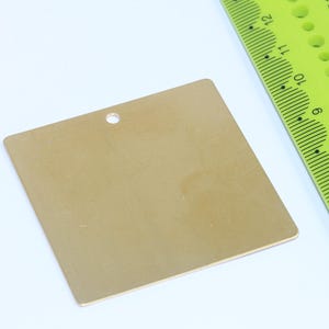 Könnte beinhalten: Ein quadratisches, goldfarbenes Etikett mit einem Loch oben, wahrscheinlich zum Aufhängen. Ein neongrünes Lineal mit schwarzen Markierungen befindet sich neben dem Etikett. Das Etikett ist etwa 7.6 cm breit.