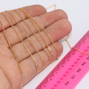 Könnte beinhalten: Eine zarte, goldfarbene Kette liegt neben einem rosa Lineal. Die Kette wird in einer Hand gehalten, und das Lineal zeigt Messungen in Zentimetern an. Die feinen Kettenglieder glänzen dezent.