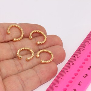 Puede incluir: Cuatro anillos de nariz de color dorado con un diseño trenzado. Los anillos están en una mano con una regla rosa en el fondo.