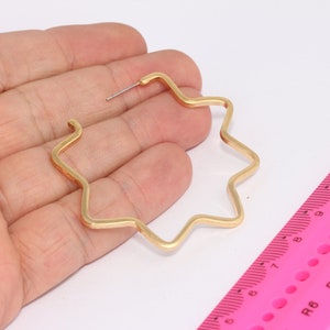 May include: A single gold-colored star-shaped hoop earring. The earring is held in a hand, with a pink ruler in the background. The earring has a post for insertion into the ear.
