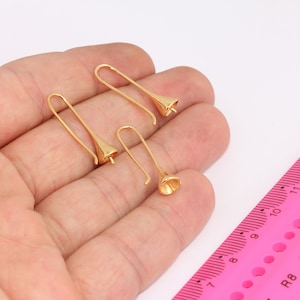 May include: Four gold-colored, bell-shaped earrings with curved ear wires are displayed. The earrings are on a hand, with a pink ruler in the background. The earrings have a delicate, floral design.