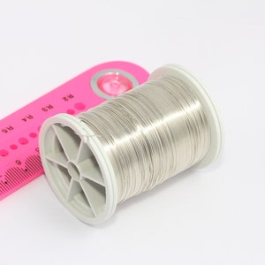 Può includere: Un rotolo di filo color argento su uno sfondo bianco. Il filo è avvolto attorno a un rotolo di plastica bianco con un righello rosa sullo sfondo.