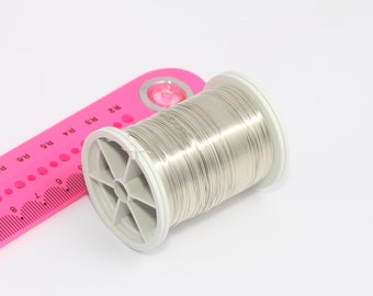 May include: A spool of silver-colored wire on a white background. The wire is wound around a white plastic spool with a pink ruler in the background.