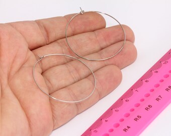 May include: Two silver hoop earrings, one larger than the other, are held in a hand against a pink ruler with black markings. The ruler measures in inches and centimeters.