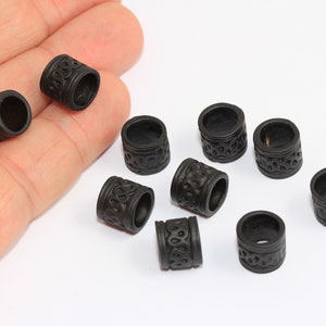 May include: Ten black hair beads with a decorative pattern. The beads are cylindrical and have a smooth, glossy finish.