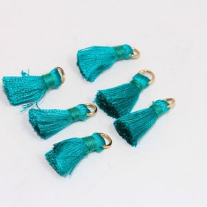 May include: Six teal blue tassel charms with gold jump rings. The tassels are made of soft, silky fabric and are approximately 1 inch in length.