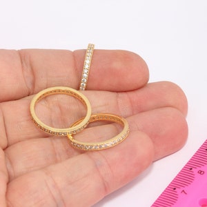 Op de afbeelding: Drie gouden ringen met een rij kleine, heldere stenen die in de band zijn gezet. De ringen liggen op een hand met een roze liniaal op de achtergrond.