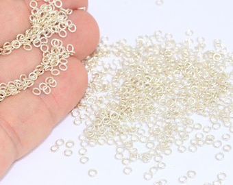 May include: A large quantity of small, silver jump rings. The rings are scattered on a white surface and a hand is holding a chain made of the same rings.