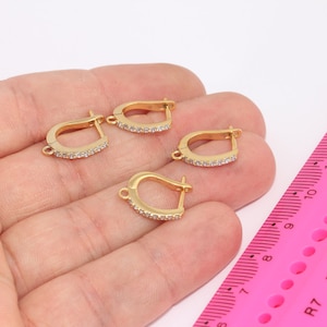 Puede incluir: Cuatro ganchos de pendientes dorados con detalles de cristal transparente. Los ganchos se muestran en una mano contra una regla rosa.