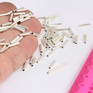 May include: A close-up of a pile of silver-colored metal tubes, some of which are arranged in a hand. A pink ruler with black markings is visible in the background.