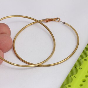May include: A pair of gold-colored hoop earrings. The earrings are circular and have a simple, classic design. The earrings are shown against a white background. A green ruler is in the background.