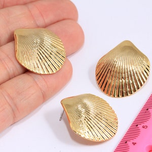 Peut inclure: Boucles d'oreilles à clous dorées en forme de coquillage. Les boucles d'oreilles ont un design texturé et strié et un fermoir à tige. Les boucles d'oreilles sont présentées sur une surface blanche.