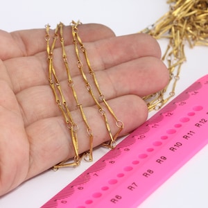 May include: A gold chain necklace with a unique design featuring alternating long and short links. The chain is laid out on a pink ruler with markings up to 15 cm.
