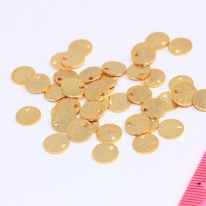 Könnte beinhalten: Ein Haufen goldfarbener runder Metallscheiben-Anhänger mit einem kleinen Loch in der Mitte. Die Anhänger sind auf einer weißen Oberfläche verstreut.