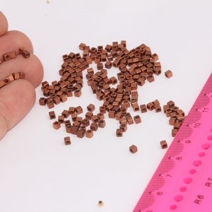 May include: A large quantity of small, copper-coloured, square-shaped beads are scattered on a white surface. A pink ruler with black markings is visible in the bottom right corner of the image. The ruler has markings for centimetres and inches.