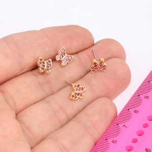 May include: Three pairs of gold butterfly earrings with pink crystal accents. The earrings are shown on a hand against a pink ruler.