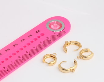 May include: Three gold-colored hoop earrings with a hinged closure. The earrings are displayed on a pink ruler with black markings.