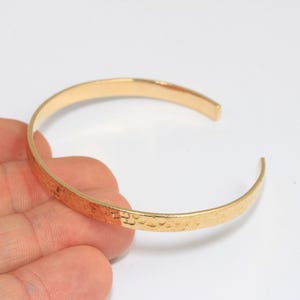 May include: A gold-toned hammered metal bangle bracelet. The bracelet is open and has a smooth, rounded edge.