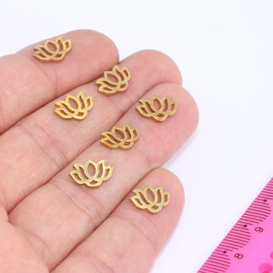 Könnte beinhalten: Sieben goldfarbene Metall-Anhänger in Form von Lotusblumen. Die Anhänger sind klein und zart und in einer Reihe auf einer Hand angeordnet.