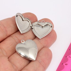 May include: Two silver heart-shaped locket pendants with a hinged opening. The lockets are shown on a hand with a pink ruler in the background.