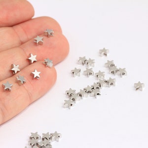 Può includere: Primo piano di una mano che tiene un mucchio di perline a forma di stella argentate. Le perline sono sparse su una superficie bianca.