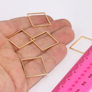 May include: Four gold-coloured metal square frames, each with a different size, are arranged on a hand. A pink ruler with black markings is in the background.