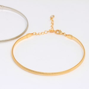 Könnte beinhalten: Zwei dünne Metallarmbänder, eines silberfarben und eines golden, mit einem zarten Kettenverschluss. Das goldene Armband hat eine strukturierte Oberfläche.
