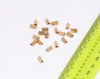 May include: A group of small, gold-colored metal triangle-shaped beads with a hole in the center. The beads are scattered on a white surface next to a yellow ruler.